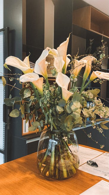 A large, elegant floral arrangement in a clear glass vase placed on a wooden table. The bouquet features creamy-white calla lilies with smooth, elongated petals and gentle curves, complemented by spri