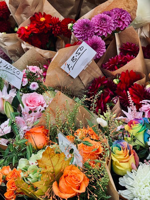 A vibrant assortment of fresh flowers arranged in multiple bouquets displayed outdoors, with some wrapped in brown craft paper and others in clear wrapping. The bouquets include a variety of blooms su
