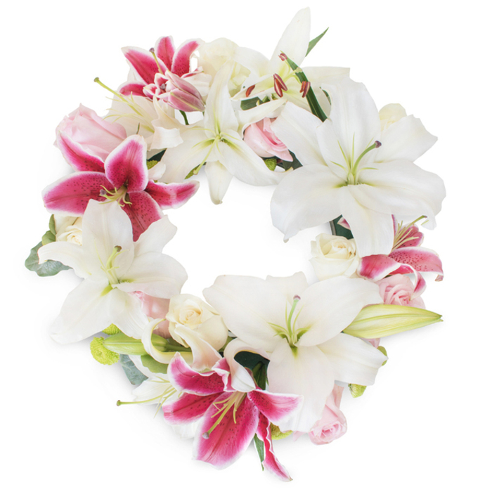 Against a pristine, luminous white background, a luxurious circular floral wreath exudes refinement and profound meaning-its expertly crafted design making it a distinguished choice for funerals or memorials in Pentonville. Prominent among the blooms are large, star-shaped lilies in snowy white, their elegant petals recalling winter light on Pentonville's historic terraces, while pale green stamens add a subtle inner glow. Punctuated throughout the design are electrifying fuchsia-pink lilies, their petals dramatically speckled and edged in deeper hues, providing eye-catching contrast and vitality. Scattered between these regal lilies are delicate, blush pink and creamy white roses, some blossoming fully with tousled, romantic petals, others in tender bud form-each lending softness and a gentle counterpoint to the bold lilies. Wisps of foliage, possibly eucalyptus or slender lily greens, peek from behind the blooms, their muted tones lending subtle structure and reminding one of the neighbourhood's hidden gardens. The wreath creates a full, unbroken circle-a symbol of remembrance and unity-its dense, harmonious composition crafted for moments of deep reflection. Such a floral tribute, imbued with both vitality and peace, is ideal for expressing sympathy, honoring loved ones, or celebrating life's enduring spirit within the close-knit Pentonville community.