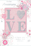 The photograph presents an anniversary card designed with the same tenderness and care that a florist might pour into an intimate, romantic arrangement. At the card's centre, the word LOVE appears in bold yet graceful raised letters, each one coated with a fine dusting of glitter in soft blush pinks, pearly whites, and a whisper of silver. This textured lettering seems to shimmer as though the light was drifting in from a nearby Pentonville window, catching on every ridge and curve. The letters rest on a gentle blush background that deepens behind them and softens toward the edges, creating a dreamy, almost ethereal glow. Surrounding this focal point, delicate botanical illustrations sweep across the surface: stylised blossoms, tiny buds, and curving leaves, sketched with the sensitivity of a florist planning an anniversary bouquet for a home near Pentonville Road or a cosy apartment close to King's Cross. The flowers are not bold or overly bright, but instead rendered in pastel pinks, soft greys, and creamy whites, with tiny silvery touches that suggest foil or dew on petals. These floral swirls move diagonally and horizontally, giving the card an easy sense of motion and improvisation, as if the flowers are drifting in a gentle breeze. Nestled among the artwork, an elegant, handwritten-style script expresses sentiments of lasting love and shared memories, the dark ink standing out gently against the soft background. The overall mood of the card is one of quiet celebration and deep affection-less like a grand display and more like a cherished keepsake, perfectly suited for an anniversary evening shared in one of Pentonville's calm, characterful homes.
